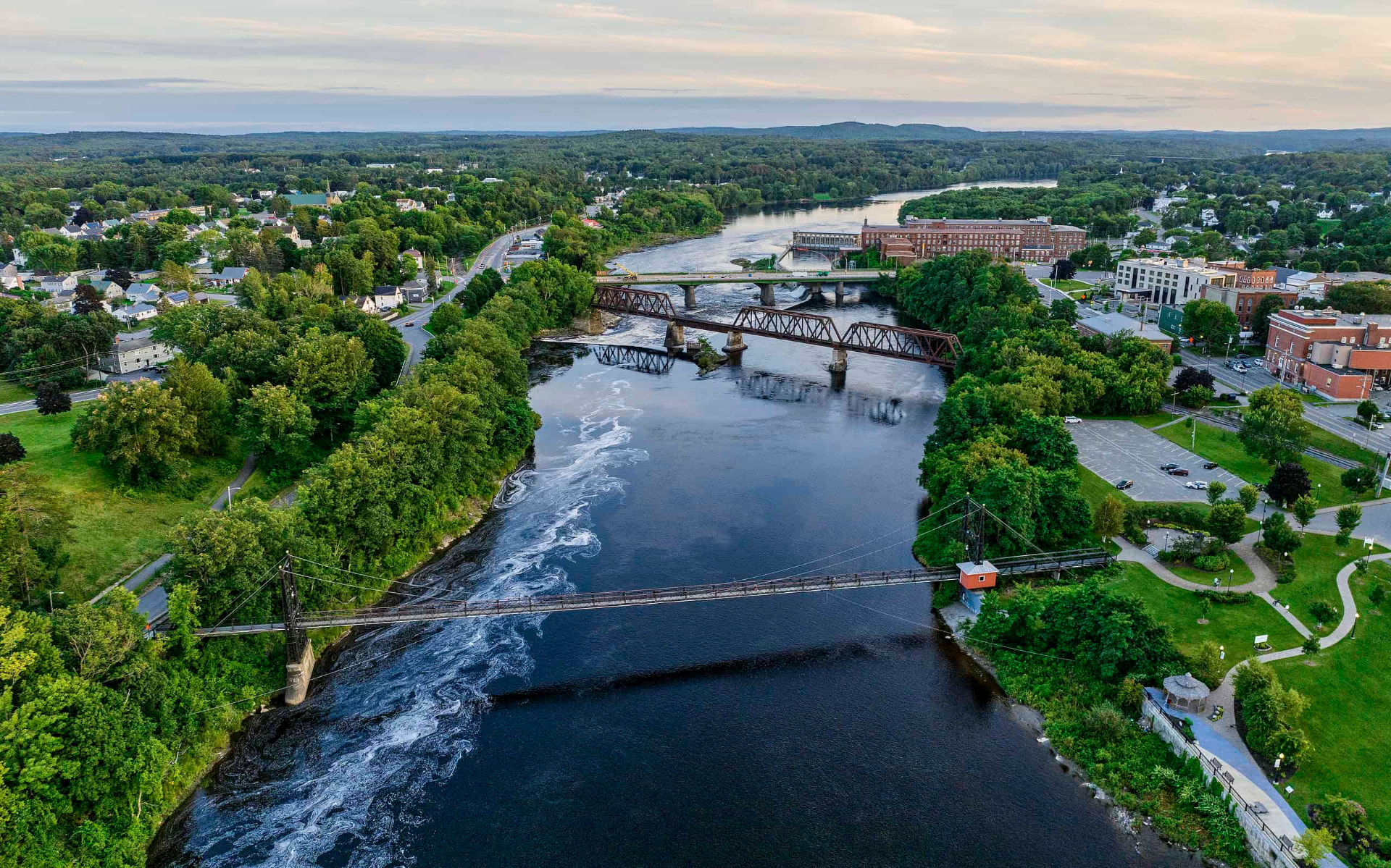 Aerial view of the Kennebec River in Maine with bridges, mills, and rolling forested hills at dusk
